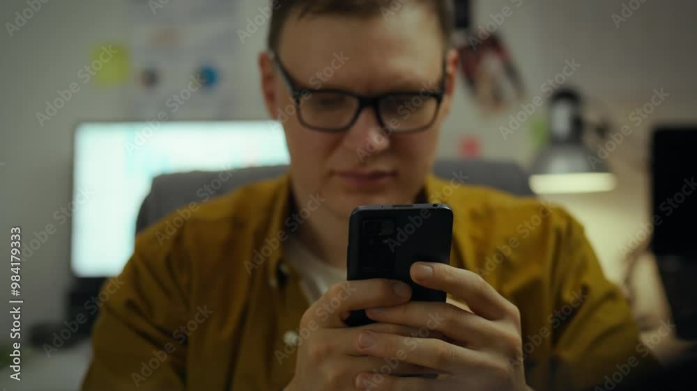 A young college student is immersed in his phone. In a dorm of a technical educational institution, a young man sits at a computer, enthusiastically looking at the screen of a smartphone A young college student is immersed in his phone. In a dorm of a technical educational institution, a young man sits at a computer, enthusiastically looking at the screen of a smartphone