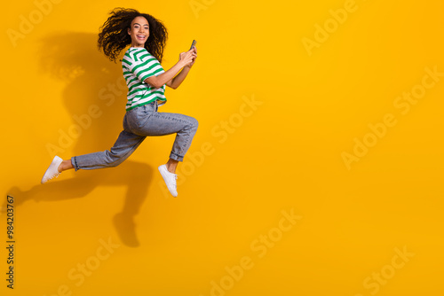 Full body portrait of nice young girl hold phone run jump empty space wear striped t-shirt isolated on yellow color background