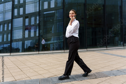 A Young Woman Walks Confidently in Professional Attire Near a Modern Glass Building on a Sunny Day