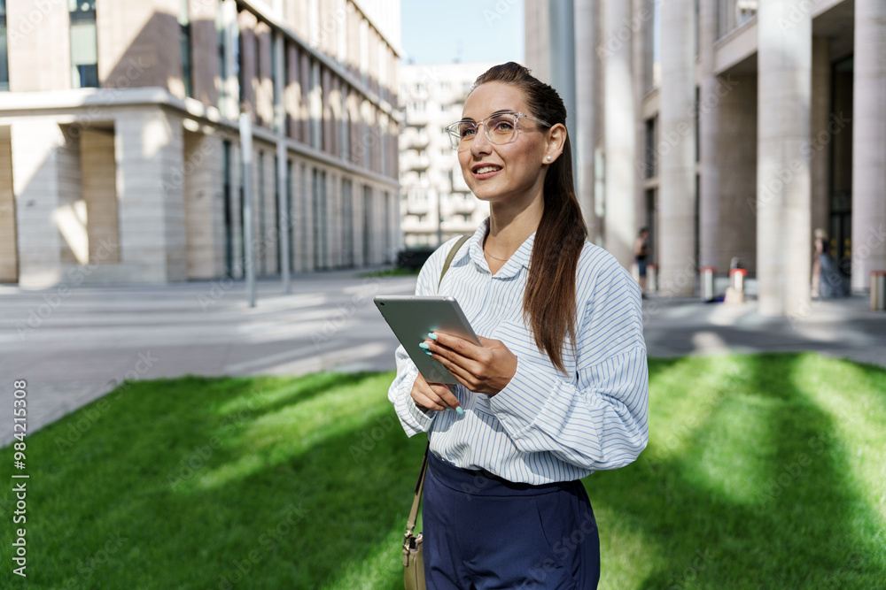 A Young Professional Stands Confidently Outdoors Holding a Tablet in a Modern Urban Setting With Sunlight Filtering Through Buildings A Young Professional Stands Confidently Outdoors Holding a Tablet in a Modern Urban Setting With Sunlight Filtering Through Buildings