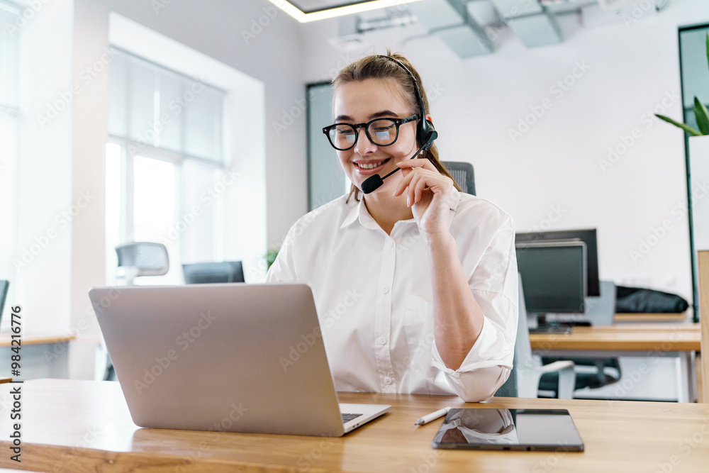 A Young Professional Wearing Glasses Engages in a Conversation While Working at a Laptop in a Bright Office Environment A Young Professional Wearing Glasses Engages in a Conversation While Working at a Laptop in a Bright Office Environment