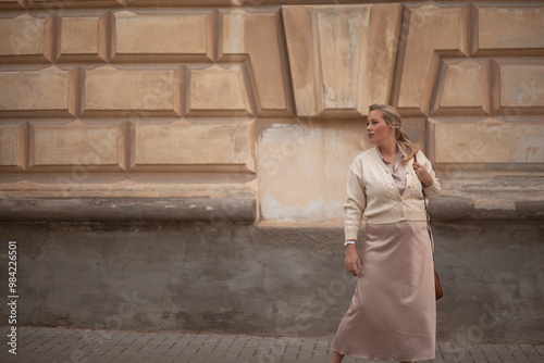 The girl plus the site is stylishly dressed in pastel clothes, a beige knitting blazer, glasses, a skirt and a silk blouse walks down the street against the background of the ancient walls of the city