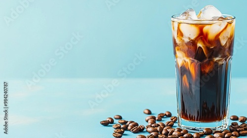 Rich and dark iced coffee in a tall glass with ice cubes, surrounded by scattered brown coffee beans on a light blue background. photo