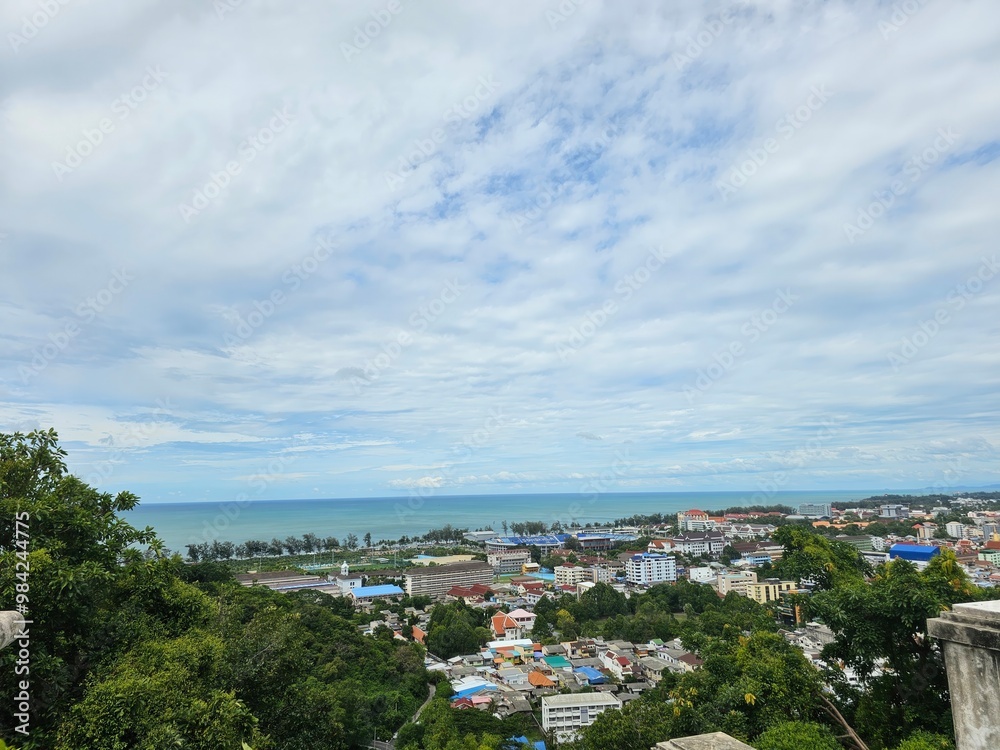 Fototapeta premium A panoramic view of a coastal town with lush greenery, ocean waves, and a cloudy sky, showcasing the vibrant roofs of houses below.