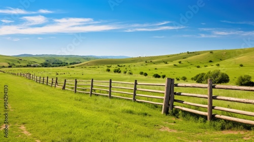 A rustic wooden fence leading through a serene countryside landscape, with rolling green hills in the distance and a clear blue sky, creating a peaceful and pastoral scene.