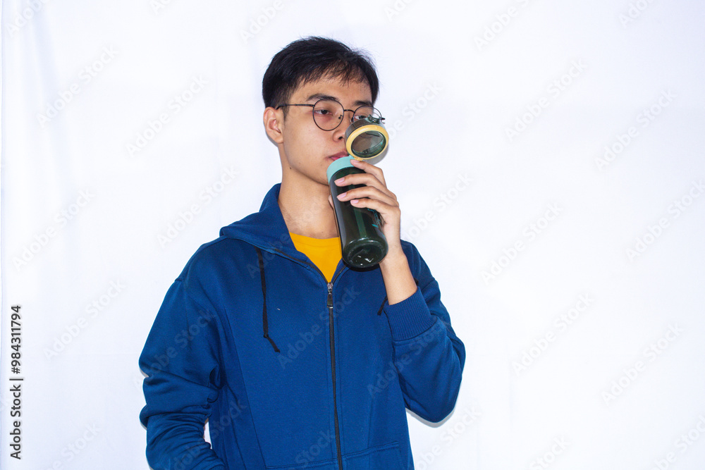 A teenager wearing a dark blue jacket and yellow shirt is relaxing while drinking water from a bottle. A teenager wearing a dark blue jacket and yellow shirt is relaxing while drinking water from a bottle.