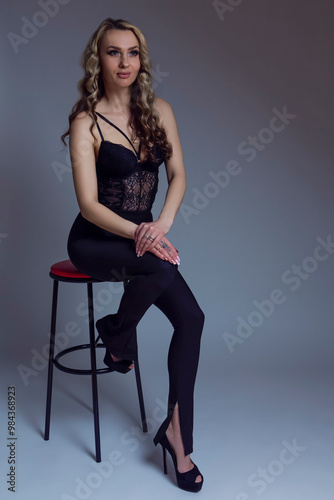 Full Length Portrait of One Smiling Positive Blond Winsome Sexy Long Curly Hair Young Caucasian Female in Black Lingerie Posing On Stool