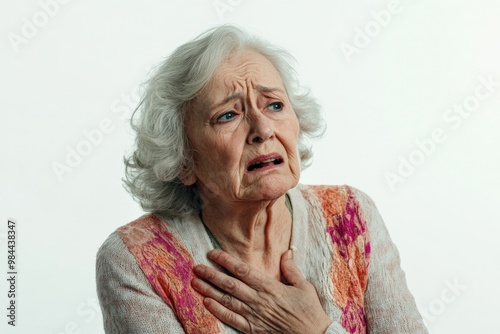 An image of a concerned elderly woman clasping her chest, dressed in a colorful cardigan, highlighting her anxious facial expression against a plain white backdrop. photo