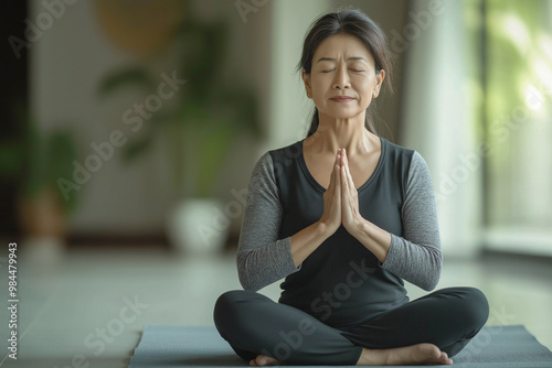 Free Download A 50-year-old Asian woman in a meditative pose in dark clothes. A woman sits on a mat with her legs crossed and her hands folded in a prayer position and meditates. The concept of calming down - GFXLoad
