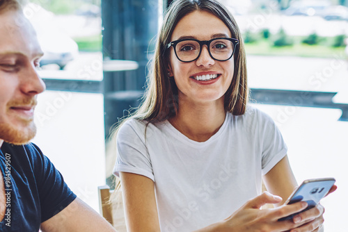 Portrait of cheerful excited hipster girl in stylish eyeglasses for vision correction looking at camera during leisure time with male friend, happy smiling hipsters using cellular gadgets for chatting Free Download Portrait of cheerful excited hipster girl in stylish eyeglasses for vision correction looking at camera during leisure time with male friend, happy smiling hipsters using cellular gadgets for chatting - GFXLoad