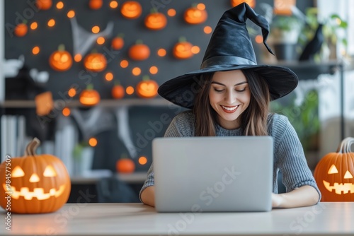 Spellbinding Work from Home: A young woman in a witch's hat smiles bewitchingly at her laptop, embracing the spirit of Halloween while working remotely amidst a festive backdrop of glowing pumpkins.  photo