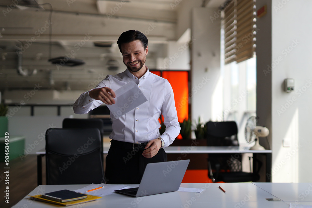 A man is standing in front of a desk with a laptop and a stack of papers. He is smiling and he is happy A man is standing in front of a desk with a laptop and a stack of papers. He is smiling and he is happy