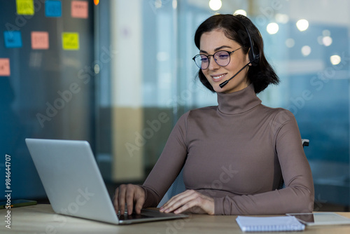 Professional woman wearing headset working on laptop in office environment. Engaged in remote work and communication, utilizing modern technology.