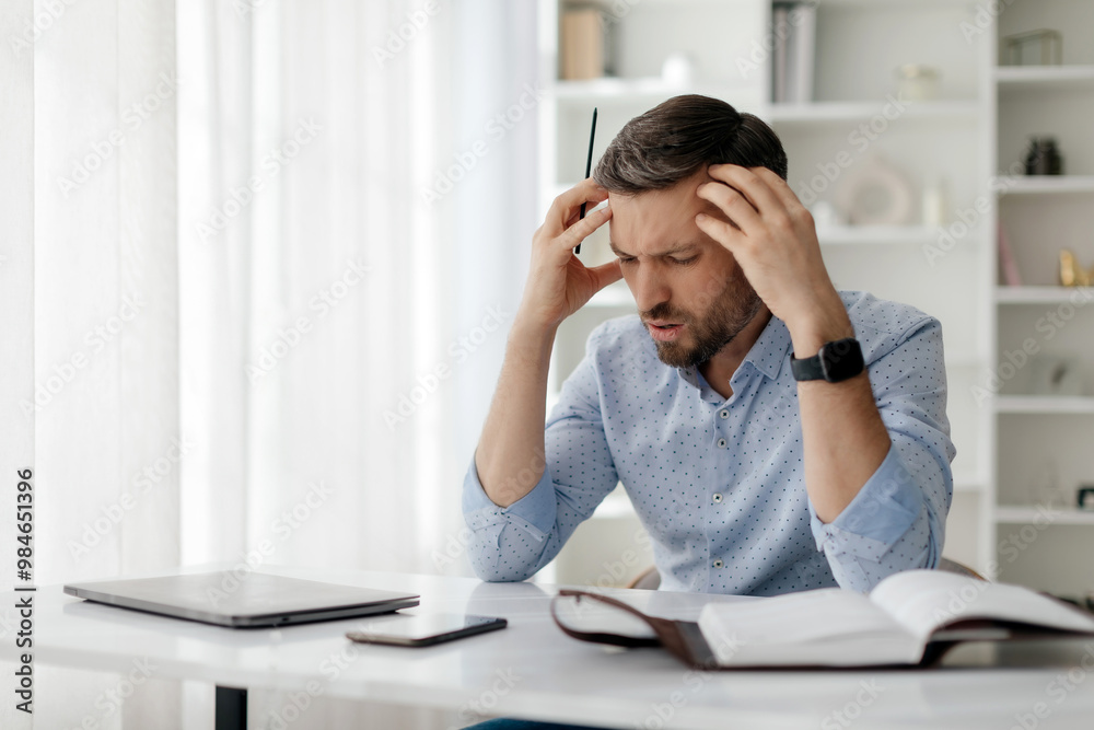 A man appears overwhelmed while sitting at a desk with a laptop, notebook, and phone, visibly stressed as he grapples with his workload during a sunny afternoon. A man appears overwhelmed while sitting at a desk with a laptop, notebook, and phone, visibly stressed as he grapples with his workload during a sunny afternoon.