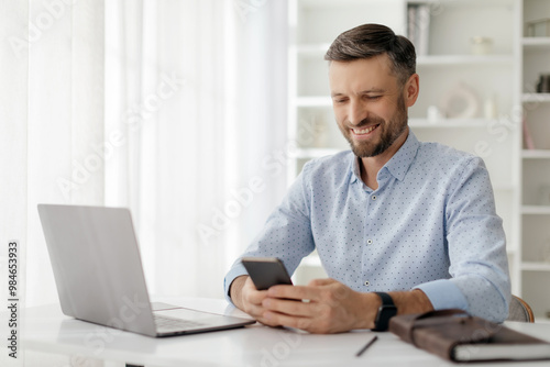 A man smiles as he interacts with his smartphone, seated at a desk with a laptop in a bright and airy office environment filled with natural light.