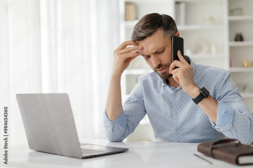 A man sits at a desk with a laptop, appearing stressed as he speaks on the phone. The bright, airy room adds to the tension of his conversation. A man sits at a desk with a laptop, appearing stressed as he speaks on the phone. The bright, airy room adds to the tension of his conversation.