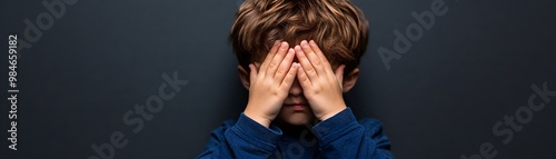 A young boy covers his eyes with his hands, conveying a sense of shyness or playfulness against a dark background.