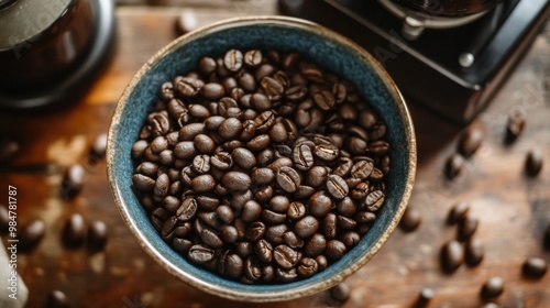 An overhead view of a bowl filled with whole coffee beans, surrounded by a few scattered beans and a coffee grinder in the background.