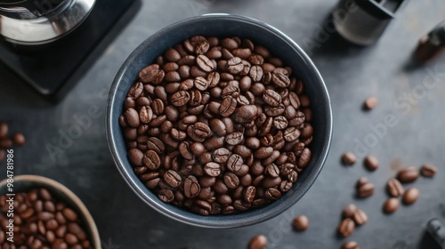 An overhead view of a bowl filled with whole coffee beans, surrounded by a few scattered beans and a coffee grinder in the background.