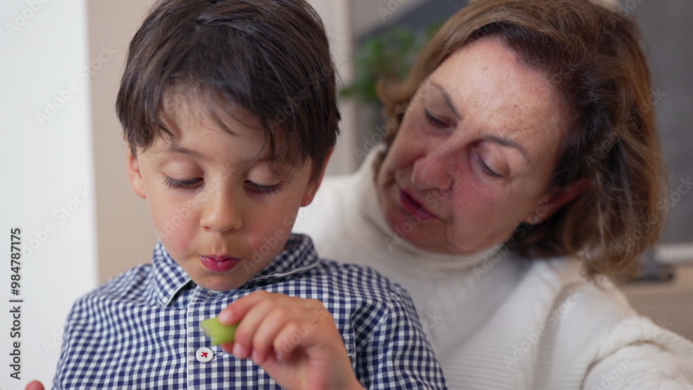 Grandmother gently feeding reluctant young boy at the table, experiencing a moment of care and connection during a family meal Grandmother gently feeding reluctant young boy at the table, experiencing a moment of care and connection during a family meal