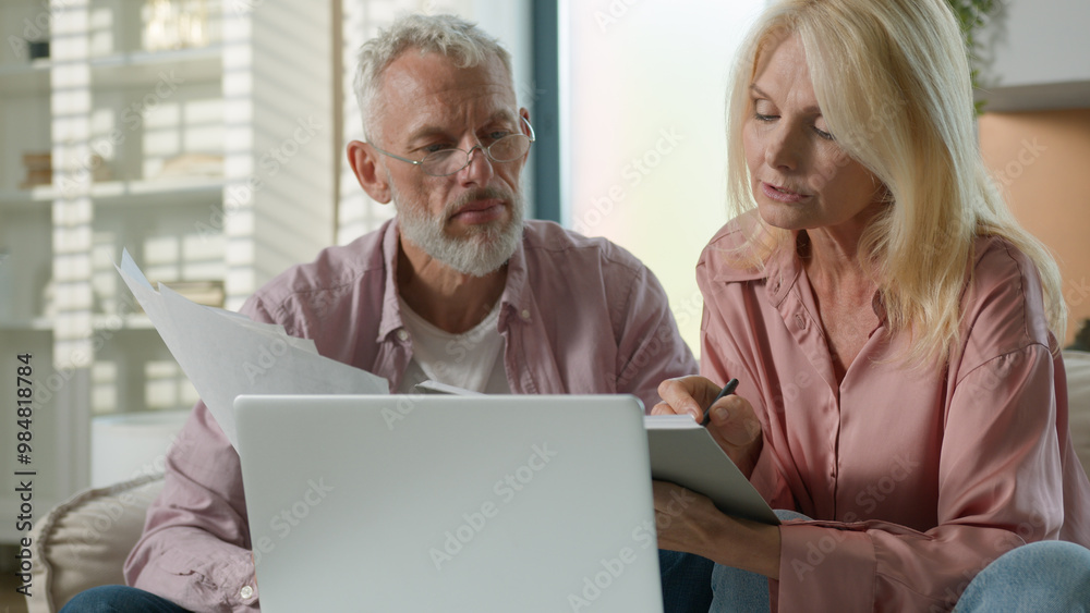 Serious senior retired elderly Caucasian couple old female wife male husband working documents financial papers use laptop computer pc. Woman man talk bills money family budget paperwork couch inside Serious senior retired elderly Caucasian couple old female wife male husband working documents financial papers use laptop computer pc. Woman man talk bills money family budget paperwork couch inside