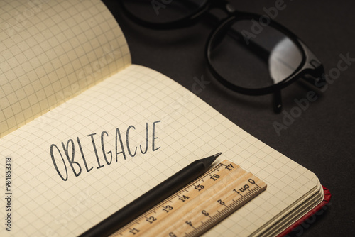 A handwritten inscription "Obligacje" on a grille of an open notebook on a wooden countertop, next to a black pencil, lighting of light. (selective focus), translation: Bonds