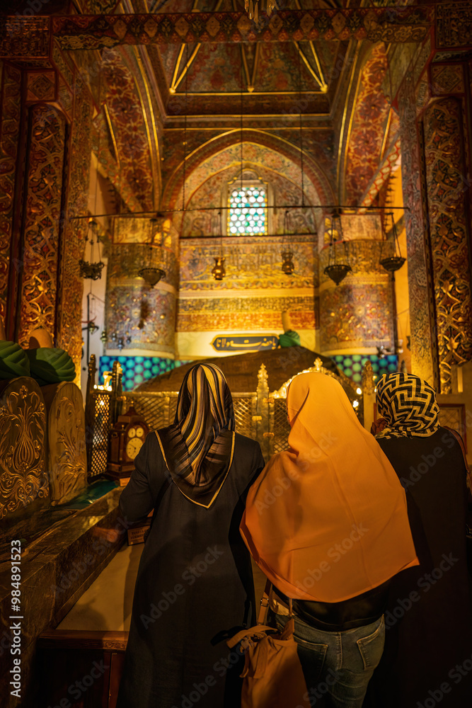 Three unidentified women wearing headscarves visit a shrine, kneeling in prayer before a decorated tomb or mosque interior