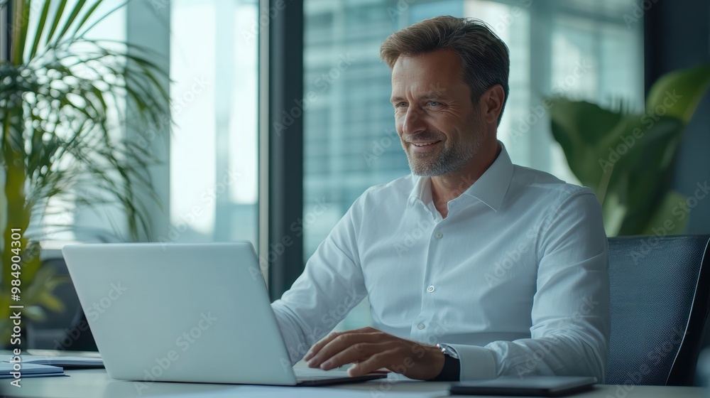 Smiling mature adult business man executive sitting at desk using laptop. Happy busy professional mid aged businessman ceo manager working on computer corporate technology in office Smiling mature adult business man executive sitting at desk using laptop. Happy busy professional mid aged businessman ceo manager working on computer corporate technology in office