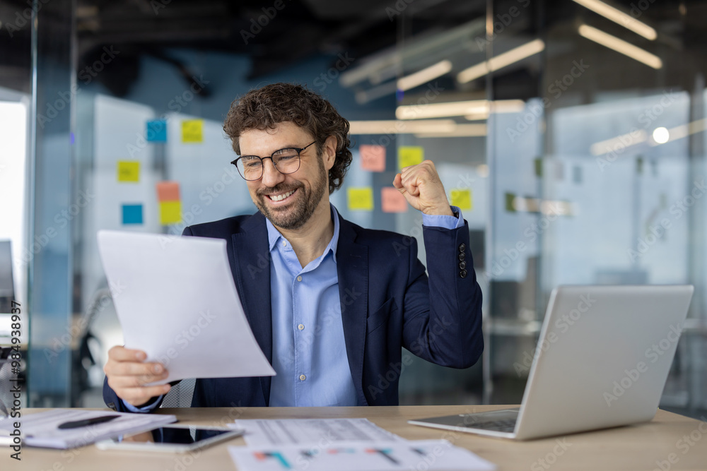 Joyful businessman in office environment showing excitement upon reading positive document. Wearing suit, surrounded by technology including laptop and phone. Demonstrates business success Joyful businessman in office environment showing excitement upon reading positive document. Wearing suit, surrounded by technology including laptop and phone. Demonstrates business success