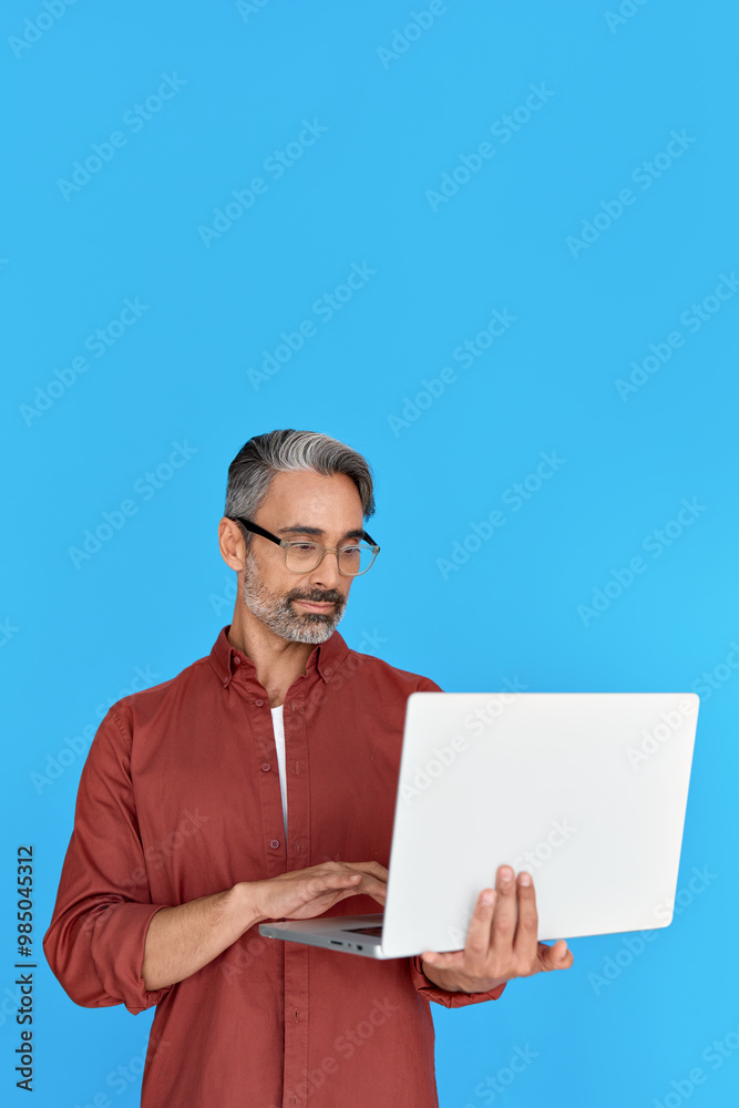 Busy mature man using laptop standing isolated on blue background. Middle aged professional businessman holding computer for work or elearning, searching job online. Vertical photo Busy mature man using laptop standing isolated on blue background. Middle aged professional businessman holding computer for work or elearning, searching job online. Vertical photo