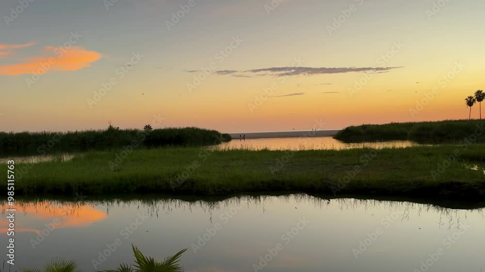 Soft pink clouds reflect in the calm waters of a coastal lagoon at dusk, creating a beautiful and serene scene. The reflection adds depth to the tranquil setting, framed by lush grass and sandy shores Soft pink clouds reflect in the calm waters of a coastal lagoon at dusk, creating a beautiful and serene scene. The reflection adds depth to the tranquil setting, framed by lush grass and sandy shores