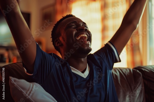 Joyful black man cheers passionately at home wearing navy blue jersey, arms raised in warm indoor with natural pouring in from window. Smiling face looks directly at camera, surrounded by cozy living photo