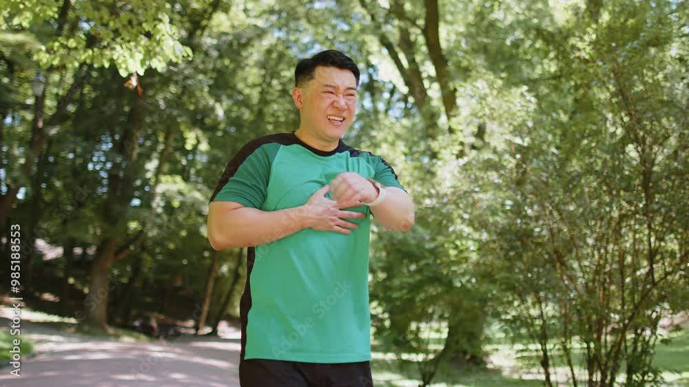 Young adult Asian runner having heart attack or chest pain after running workout in park on sunny summer day. Tired Japanese athlete suffers from tension in the heart. Health care and sports concept.