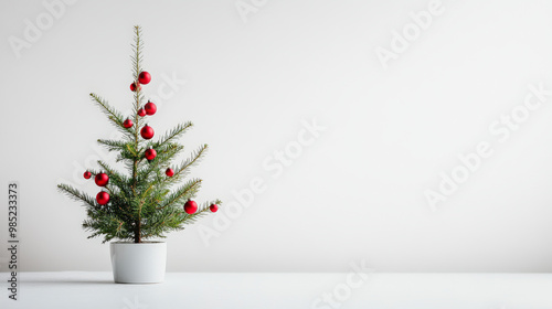 A small potted Christmas tree decorated with red baubles, placed on a plain white surface against a minimalist white background, creating a festive and simple scene.