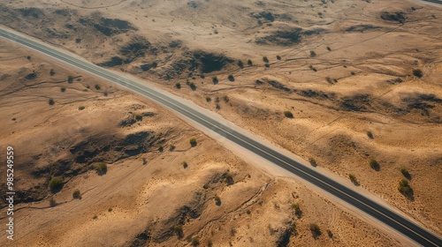 Aerial view of a desert road winding through a dry, sandy terrain. A rural path with a stone structure in the distance. The landscape is dominated by brown hills and a sunny sky.