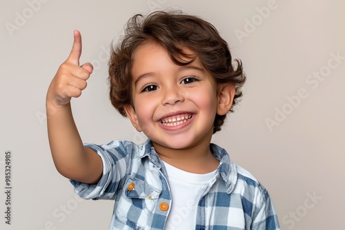 Latin kid wearing casual clothes smiling cheerfully points with finger upwards. Happy boy gestures to something off-camera. Kid confident expression, cheerful smile convey sense of welcome,