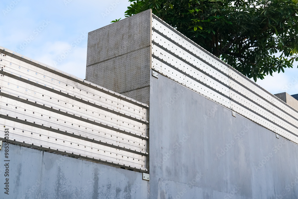 Modern concrete wall with metal panel accents, clean geometric lines, and industrial aesthetics, set against a blue sky with green foliage peeking from behind.