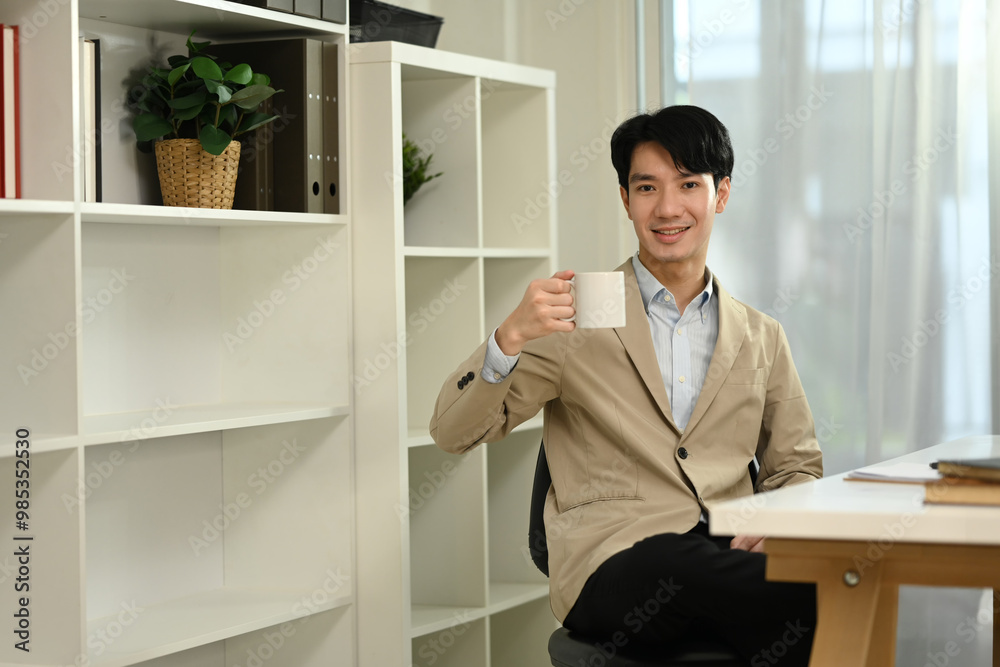 Fototapeta premium Positive young entrepreneur in suit drinking hot coffee at modern office