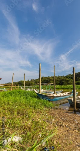 Vertical 4k time lapse on a rain forest river. Tourist and fisher men boats on the bank. Veil clouds on the blue sky. Small boat floats in river water. The wild untouched jungle in the background. 