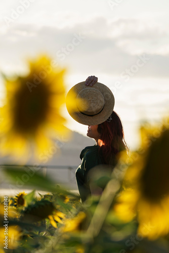 A smiling middle-aged woman standing in a sunflower field at sunset. The individual is wearing a hat and has long hair