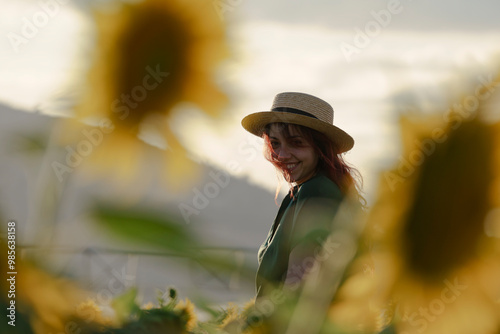 A smiling middle-aged woman standing in a sunflower field at sunset. The individual is wearing a hat and has long hair