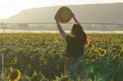 A middle-edged woman standing in a sunflower field during the golden hour. They are seen from behind, holding a straw hat up in the air with both hands.