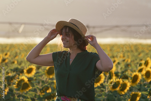 A smiling middle-aged woman standing in a sunflower field at sunset. The individual is wearing a hat and has long hair