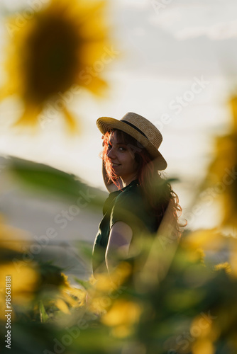 A smiling middle-aged woman standing in a sunflower field at sunset. The individual is wearing a hat and has long hair