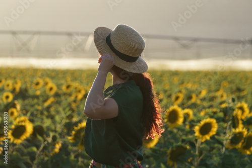 A smiling middle-aged woman standing in a sunflower field at sunset. The individual is wearing a hat and has long hair