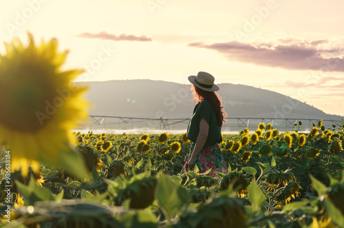 A smiling middle-aged woman standing in a sunflower field at sunset. The individual is wearing a hat and has long hair