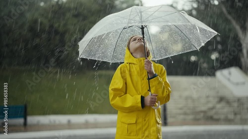 Happy teenage girl in a yellow jacket holds an umbrella and watches water drops flow down it during heavy rain while walking in the park