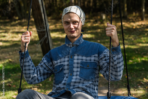 A cheerful young guy riding a swing in the village. A guy in a shirt and a scarf photo