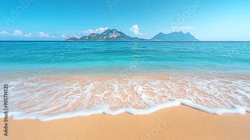 A calm ocean with a white foamy wave lapping at a sandy shore.  A mountainous island sits in the distance on a clear blue day.