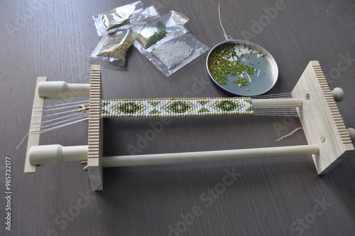 A close-up of hands crafting a beaded bracelet on a specialized loom. The bracelet features a traditional pattern with colorful beads. Tools and materials are neatly arranged, showcasing precise